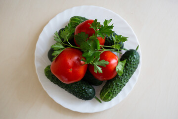 A top view of fresh tomatoes, sliced cucumbers and sprig of parsley on the white plate on the light table. Seasoning vegetables on a table. Healthy ingredient for salad.