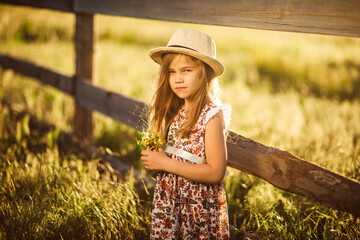 girl in hat standing next to fence in village with bouquet of wild flowers