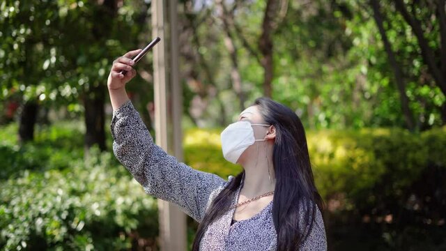 Chinese girl taking selfie outside with phone wearing mask. Masked China woman outdoors takes photos cell phone, posing for the camera. Cute girl with face mask to protect from virus and pollution.