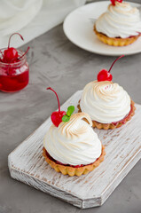 Tartlets with cherry filling and Italian meringue with a cocktail cherry on top on a board on a concrete background. Vertical orientation. Copy space. Food background.