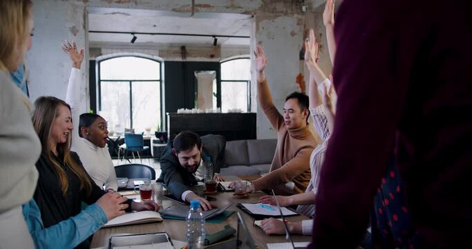 Happy business people team put hands together, funny Caucasian boss falls on table at loft office meeting slow motion.