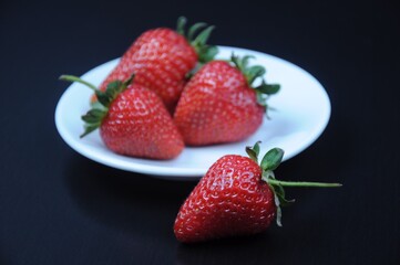 A large strawberry in front of a white saucer and three ripe red juicy strawberries on it on a black background