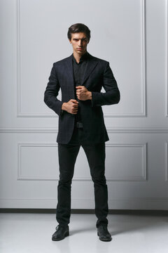 Vertical view. Full length picture of an elegant young man adjusting his suit while looking to his side, looking at camera, on white background.