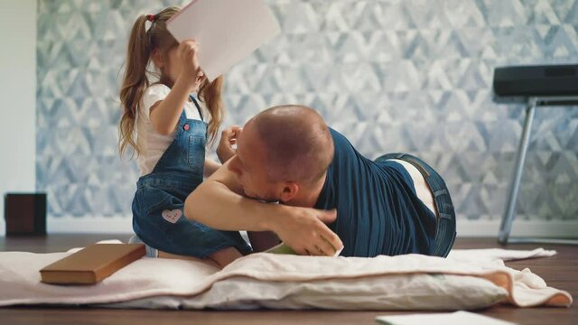 playful little girl with ponytails hits shaved bald father with notebook lying on floor with soft blanket in light room at quarantine time