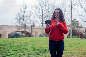 Portrait of happy pretty caucasian young woman model showing chocolate  pointing the piece with index finger, in the park, orange sweater and jeans, long curly hair. Place for your text in copy space.