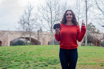Portrait of happy attractive caucasian young woman holding chocolate, keeping finger crossed, in the park, orange sweater and jeans, long curly hair. Place for your text in copy space.