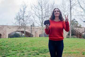 attractive caucasian young woman model with chocolate  making victory or peace sign, in the park, orange sweater and jeans, long curly hair. Place for your text in copy space.