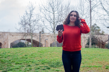 Portrait of attractive caucasian young woman holding chocolate  thinks with finger on head, in the park, orange sweater and jeans, long curly hair. Place for your text in copy space.