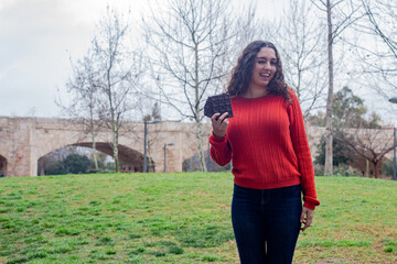 Portrait of attractive caucasian young woman model biting chocolate  winking, in the park, orange sweater and jeans, long curly hair. Place for your text in copy space.