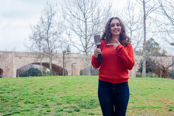 Portrait of pretty caucasian young woman showing rotating and styling brush  pointing the piece with index finger, in the park, orange sweater and jeans, long curly hair. Place for your text in copy s