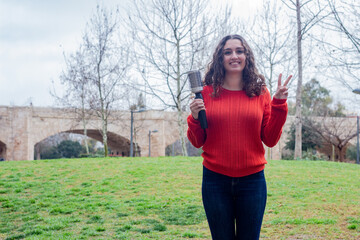 Portrait of young woman with rotating and styling brush  making victory or peace sign, in the park, orange sweater and jeans. Place for your text in copy space.