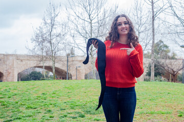 Portrait of pretty caucasian young woman model showing electronic abdominal appliance  pointing the piece with index finger, in the park, orange sweater and jeans, long curly hair. Place for your text
