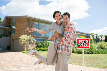 Portrait enthusiastic couple hugging outside house with For Sale sign