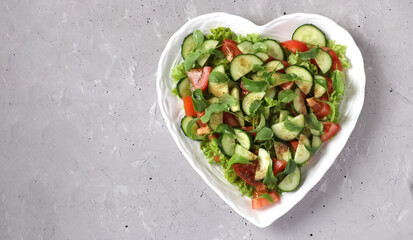 Heart-shaped plate with healthy salad with tomatoes, cucumbers, arugula and microgreens on gray concrete background, Space for text, Top view