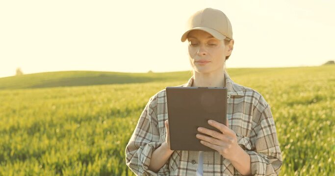 Beautiful young female farmer in hat standing in field and using tablet device. Pretty Caucasian woman tapping and scrolling on tablet computer in green margin in summer.