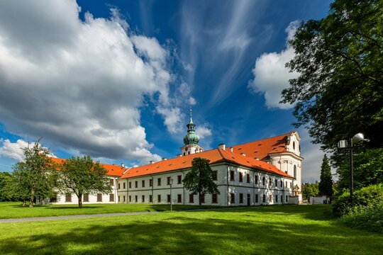 Brevnov, Prague / Czech Republic - June 11 2020: View of the first male monastery in Bohemia standing in a park with green trees on a sunny summer day, blue sky with white clouds.