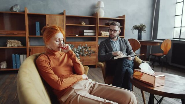 Zoom in shot of depressed middle-aged redhead woman sitting on couch and complaining to professional male psychologist during consultation in counseling office