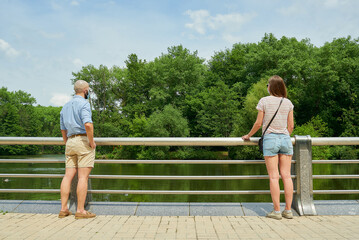 A man and a woman enjoying the view of the river keeping distance a few meters to avoid the spread of coronavirus. A guy and a girl in face masks resting leaning against the fence on the promenade