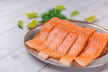 Raw vibrant salmon fillet in a serving plate with herbs, creamy sauce made from butter, garlic, and vegetables. Salmon with wedge lime on the side on a white wooden table.