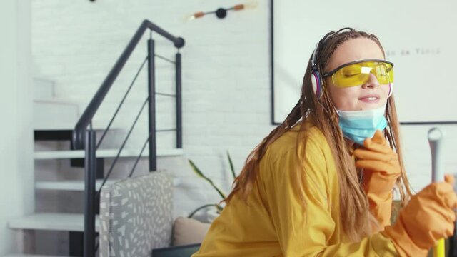 Happy young woman with mask and glasses singing and dancing with mop during clean-up enjoying music in headphones housewife fun beautiful housework cleaning slow motion