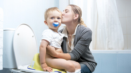 Smiling young mother teaching her toddler son using adult toilet