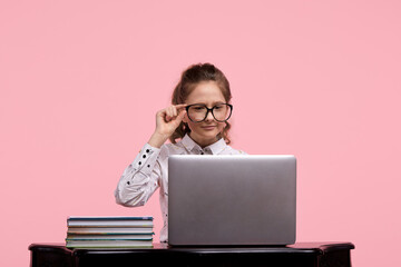 young girl sits at a table with a laptop and holds glasses with one hand.