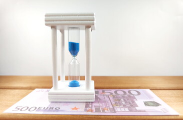 Euro coins, bundles of cash bills and an hourglass with blue sand, on a wooden background, the concept of business development from time to time. With copy space.