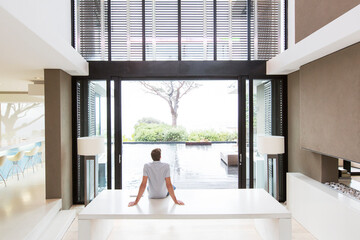 Rear view of man sitting on table looking at swimming pool through open patio door