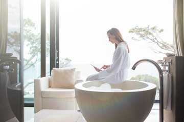 Woman wearing white bathrobe sitting on edge of modern bathtub using digital tablet