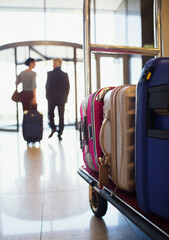Rear view of man woman leaving hotel lobby, suitcases on luggage cart in foreground - obrazy, fototapety, plakaty