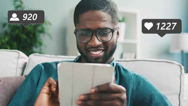 Handsome African American young man in glasses sitting on sofa at home, watching posts and liking with hearts on tablet device. Male blogger using computer and getting reactions and likes, followers.