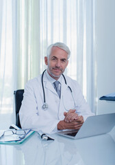 Portrait smiling mature doctor sitting at desk with laptop in office