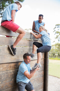 Teammates Helping Woman Over Wall On Boot Camp Obstacle Course