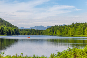 people are boating on a lake surrounded by green forests and mountains. The blue cloudy sky in the background.