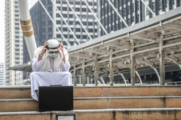 Arabic business man sad serious from coronavirus crisis. Muslim guy hopeless sitting on public ladder and stress hand on head with face mask for protection outbreak of covid-19 on cityscape background