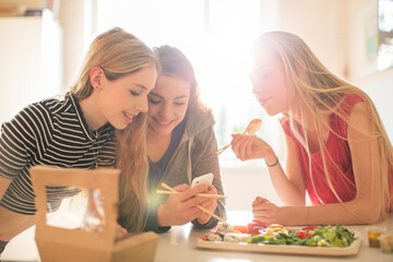 Teenage girls eating sushi and texting with cell phone in sunny kitchen