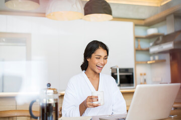 Laughing woman in bathrobe drinking coffee and using laptop