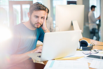 Serious creative businessman using laptop with head in hands in office