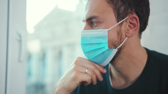 Face man wearing medical mask boring at home sitting on windowsill and looking at street on quarantine coronavirus self-isolation lockdown stress depression window boring slow motion