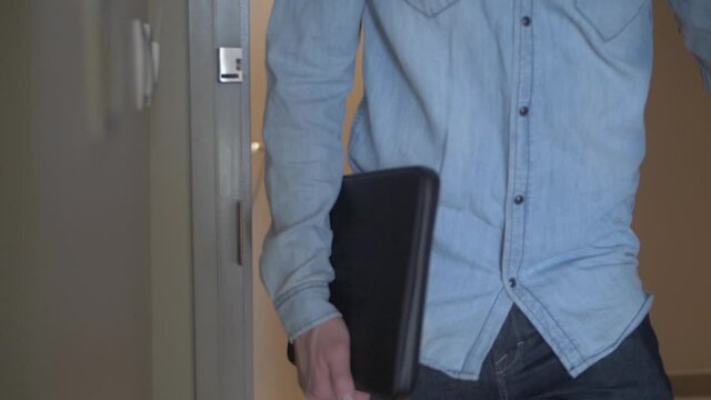 A middle-aged man in a blue shirt and jeans is walking into a hotel room carrying his laptop case. Slow motion.