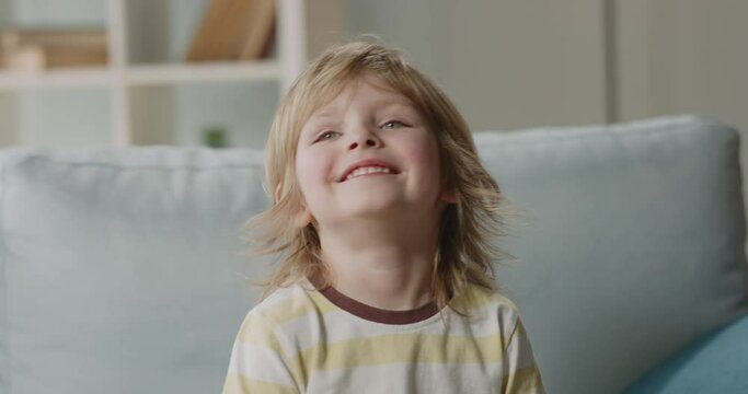 Funny little cute caucasian boy sitting on couch at home, looks at camera and positively happily smiles - real people, happy childhood concept close up 4k portrait footage