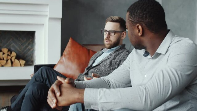 Panning Shot Of African American And Caucasian Boyfriends Sitting On Sofa Near Fireplace And Having Discussion With Female Psychologist During Couple Counseling Therapy