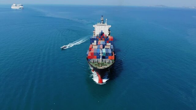 Aerial in front of beautiful cargo ship carrying container and running with pilot boat sailing out for export goods from cargo yard port to custom ocean concept freight shipping by ship forward mast