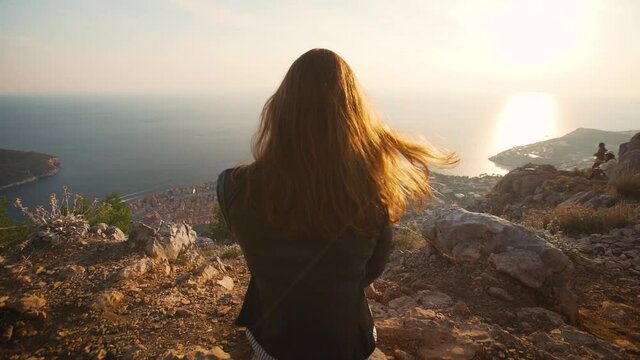 Redhead woman looking out at beautiful cliffside coastline town view in Dubrovnik Croatia at sunset push in slow motion, hair blowing in the wind.