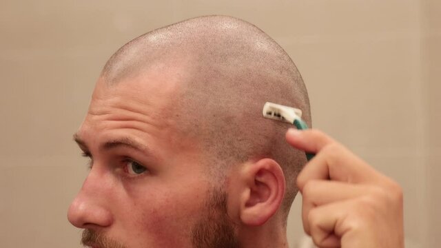 Close up of young man shaving his own bald scalp with disposable razor inside bathroom