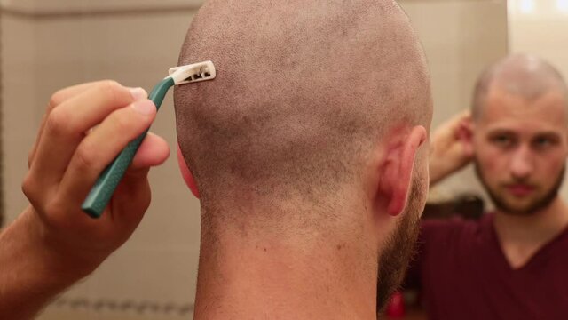 Young bald man shaving the back of his head with disposable razor blade while facing a bathroom mirror