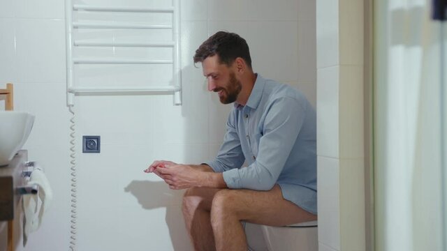 Smiling businessman speak on phone sitting on the toilet holds toilet paper mobile restroom smartphone internet home bathroom close up slow motion