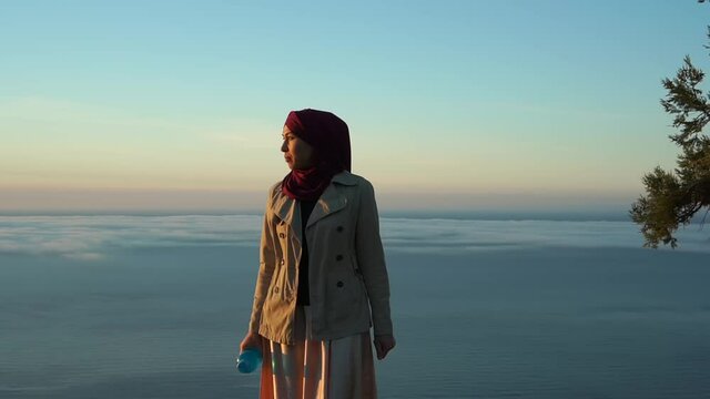 Portrait of an authentic muslim woman in a hijab with a reusable water bottle. Nature, sea, morning, landscape, sunrise
