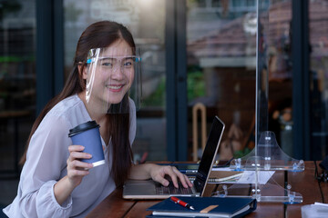 Asian working woman wearing face  shield and smiling at camera in coffeeshop, with table shield partition to protect infection from coronavirus covid-19 on table, social distancing concept