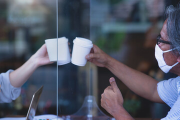 Asian man and woman click to celebrate cup of coffee after successful business deal, with table shield partition to protect infection from coronavirus covid-19 on table, social distancing concept